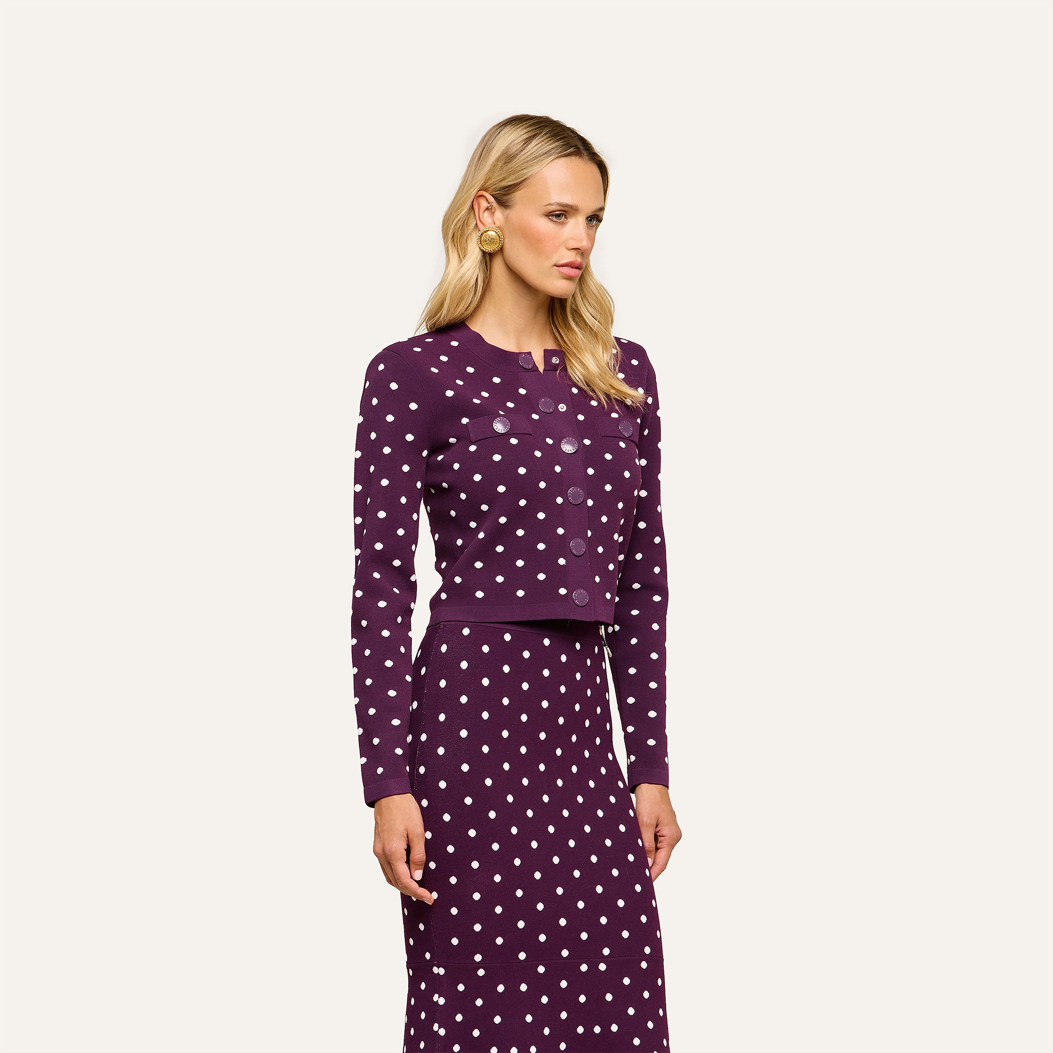 A woman with long blonde hair and gold earrings stands against an off-white background, wearing the Kota Cardigan Top—a dark purple cropped cardigan and skirt set with white polka dots—while looking slightly to the side.