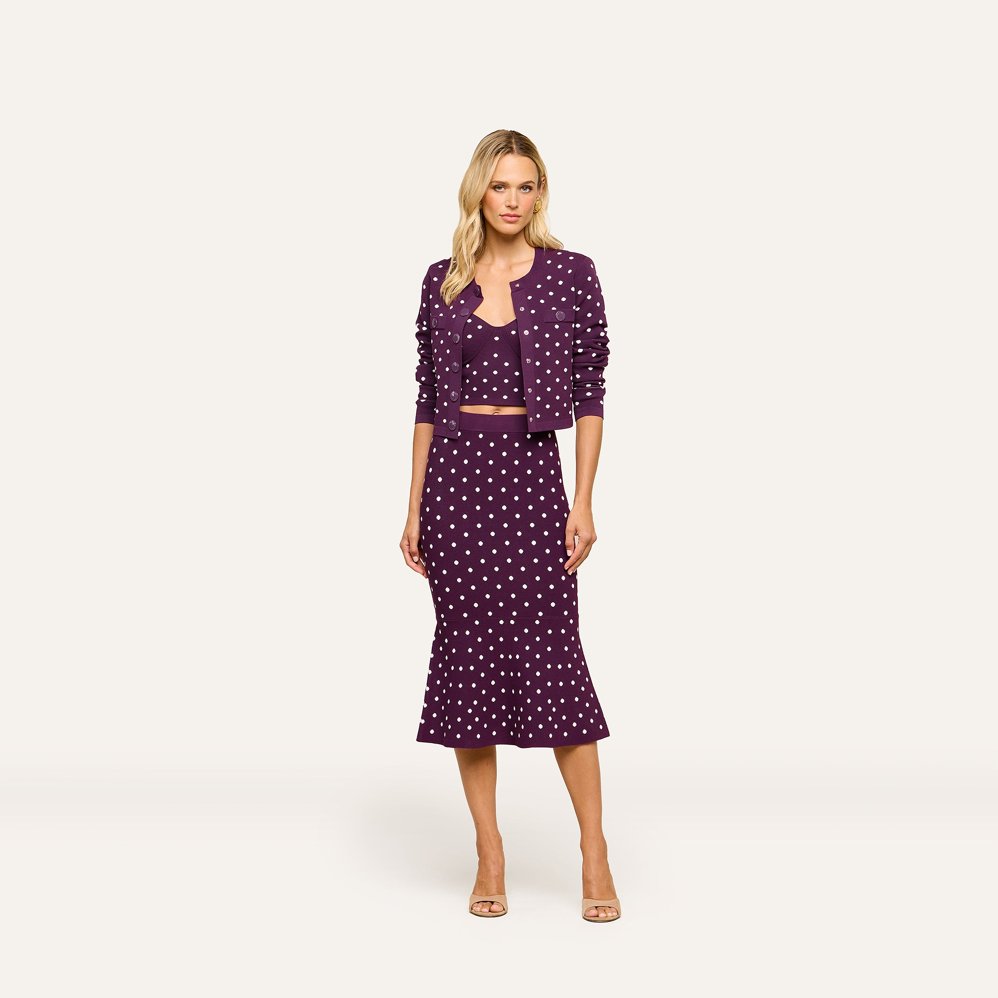 Woman wearing the Kota Cardigan Top in dark purple over a purple polka dot crop top, paired with a midi skirt and nude heels, standing against a plain white background.