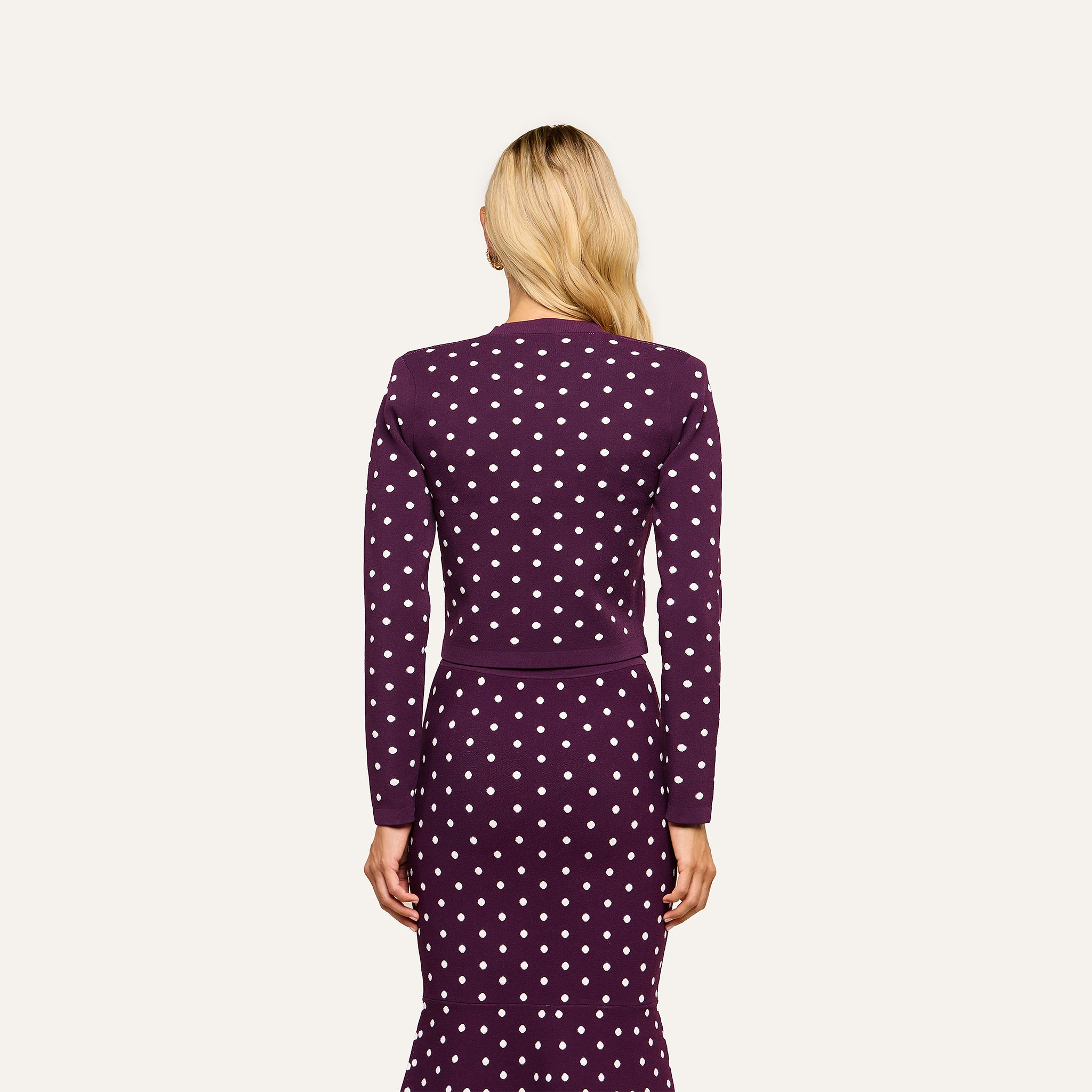 A person with long blonde hair, seen from behind, is wearing the Kota Cardigan Top in dark purple and a skirt with white polka dots. The background is plain and light-colored.