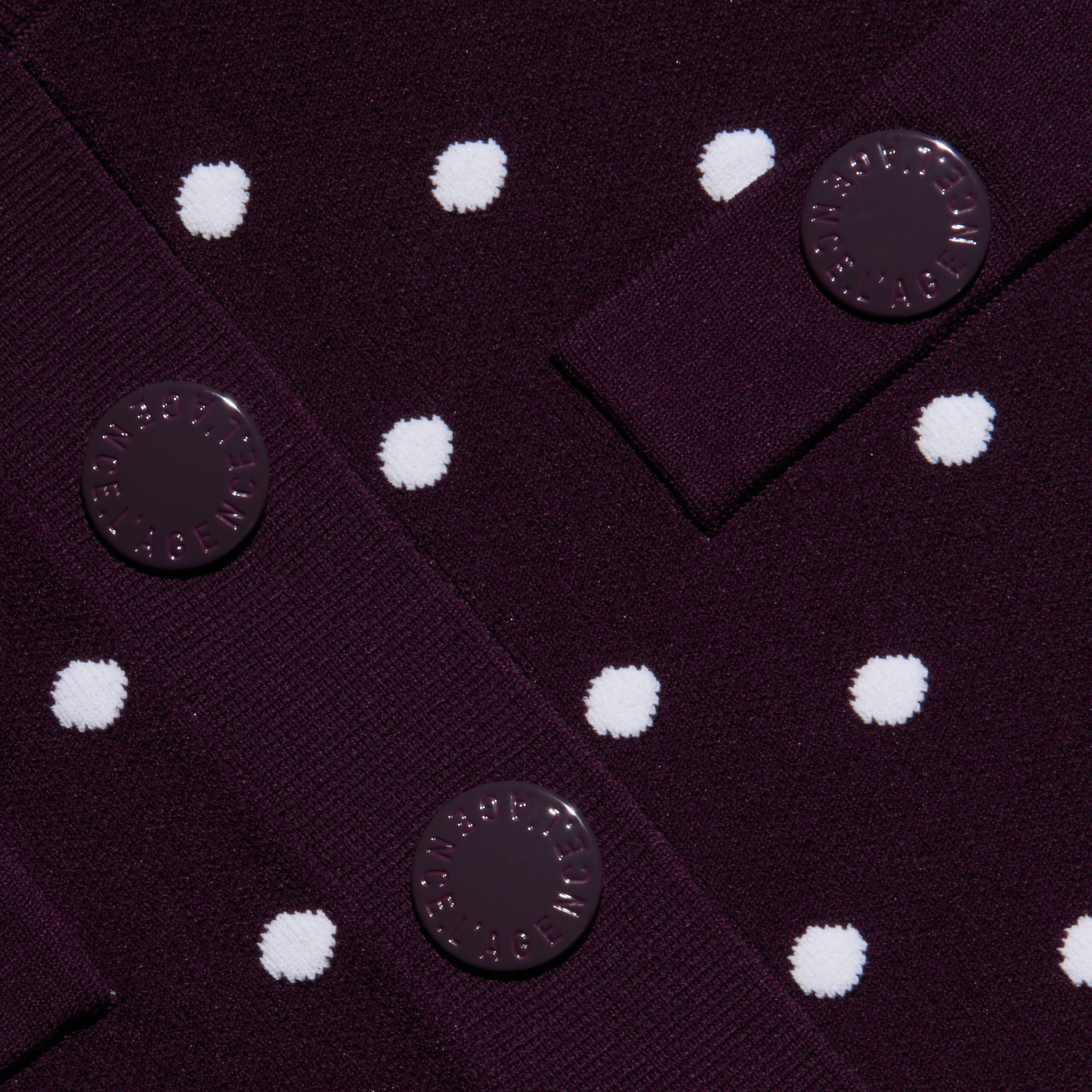 Close-up of the Kota Cardigan Top in dark purple fabric with white polka dots, featuring three round buttons with embossed text sewn onto overlapping fabric panels.
