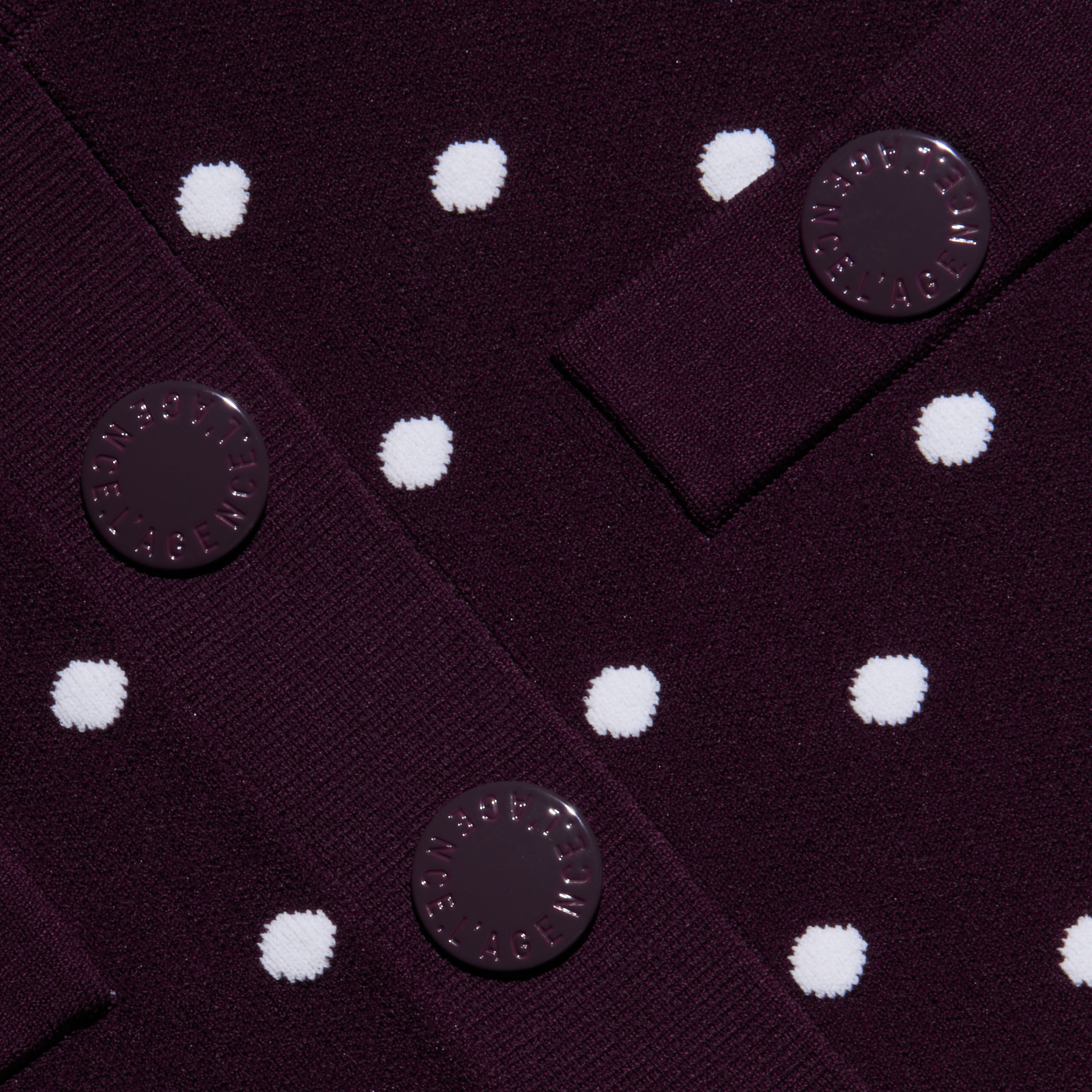 Close-up of the Kota Cardigan Top in dark purple fabric with white polka dots, featuring three round buttons with embossed text sewn onto overlapping fabric panels.
