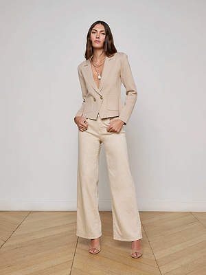 A woman with long brown hair and layered necklaces faces the camera in a neutral-toned room, wearing L'AGENCE's Sofia Knit Blazer, wide-leg cream trousers, and heeled sandals, with her hands in her pockets.