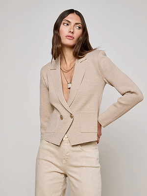 A woman models the Sofia Knit Blazer by L'AGENCE, featuring gold buttons, paired with cream pants. She stands against a plain background, wearing layered necklaces and has long brown hair, gazing to the side with a neutral expression.