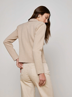 A woman with long brown hair stands facing away, wearing the Sofia Knit Blazer by L'AGENCE in metallic khaki and light-colored pants against a plain light background.