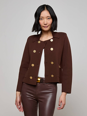 A woman with chin-length black hair wears the L'AGENCE Madilyn Knit Jacket, a tailored brown double-breasted piece with gold buttons, over a white top and brown leather pants, standing against a plain light background.
