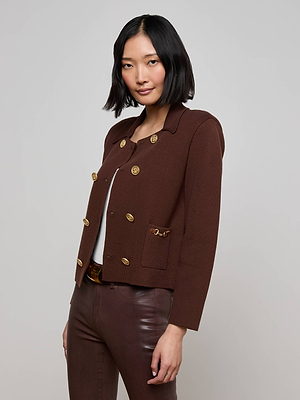 A woman with a bob haircut models the Madilyn Knit Jacket by L'AGENCE—a brown double-breasted blazer with gold buttons—styled over a white top and brown pants, standing against a plain light background and gazing confidently at the camera.