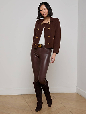 A woman with a bob haircut poses indoors against a white wall, wearing the L'AGENCE Madilyn Knit Jacket with gold buttons, a white top, brown leather pants with a belt, and dark brown knee-high boots. One hand rests on her hip.
