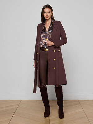 A woman stands against a light gray wall wearing the Isabel Knit Coat with gold buttons, a patterned blouse, dark brown pants, and knee-high boots. The floor is light wood in a herringbone pattern.