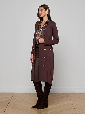 A woman wears the Isabel Knit Coat, a long maroon double-breasted piece with gold buttons, paired with a patterned blouse, matching skirt, and knee-high boots. She stands on light wood flooring against a plain white wall.