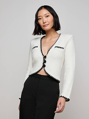A woman with chin-length black hair wears the Anaya Knit Cardigan with crochet trim and black buttons, paired with high-waisted black pants, standing against a plain light gray background.