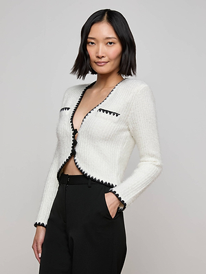 A woman with short black hair models the Anaya Knit Cardigan, which has an eyelash knit texture and bold black crochet trim, styled with black pants as she poses confidently against a plain light gray background.