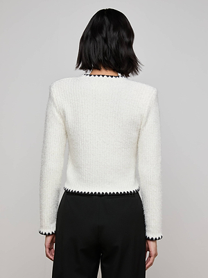 A person with short, dark hair is seen from the back wearing the Anaya Knit Cardigan, a textured white long-sleeve top with black crochet trim on the collar, cuffs, and hem, paired with black pants against a plain background.