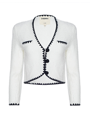 The Anaya Knit Cardigan is a white textured piece with black crochet trim on the edges, cuffs, and pockets. It features a V-neckline, long sleeves, and three black front buttons. The label reads LAGENCE.