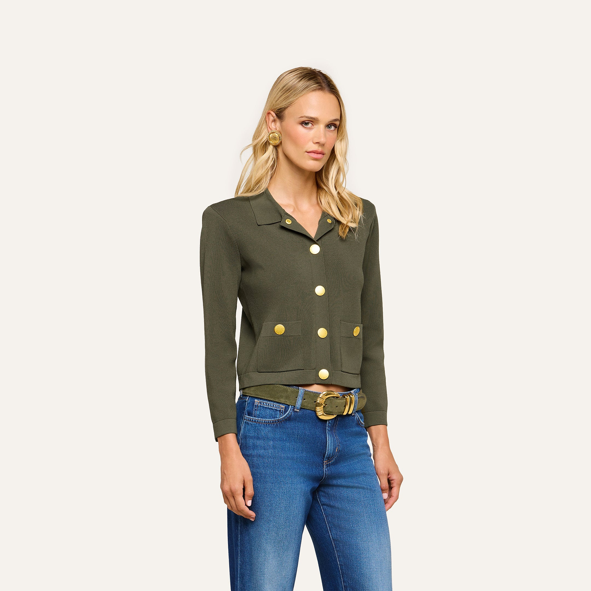 A woman wearing an Osiris Cardigan in dark green crepe knit with large gold buttons, paired with blue jeans and a gold-buckled belt, stands against a plain, light background.