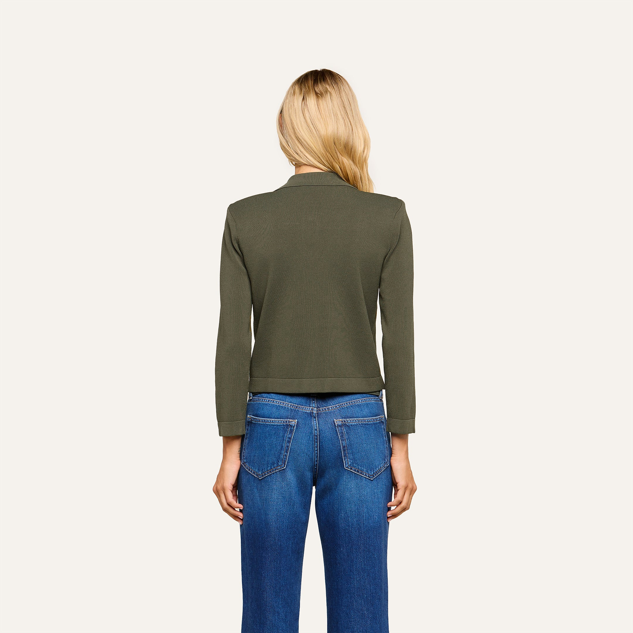 A woman with long blonde hair stands facing away, wearing the Osiris Cardigan in dark green with gold buttons and blue jeans against a plain light background.