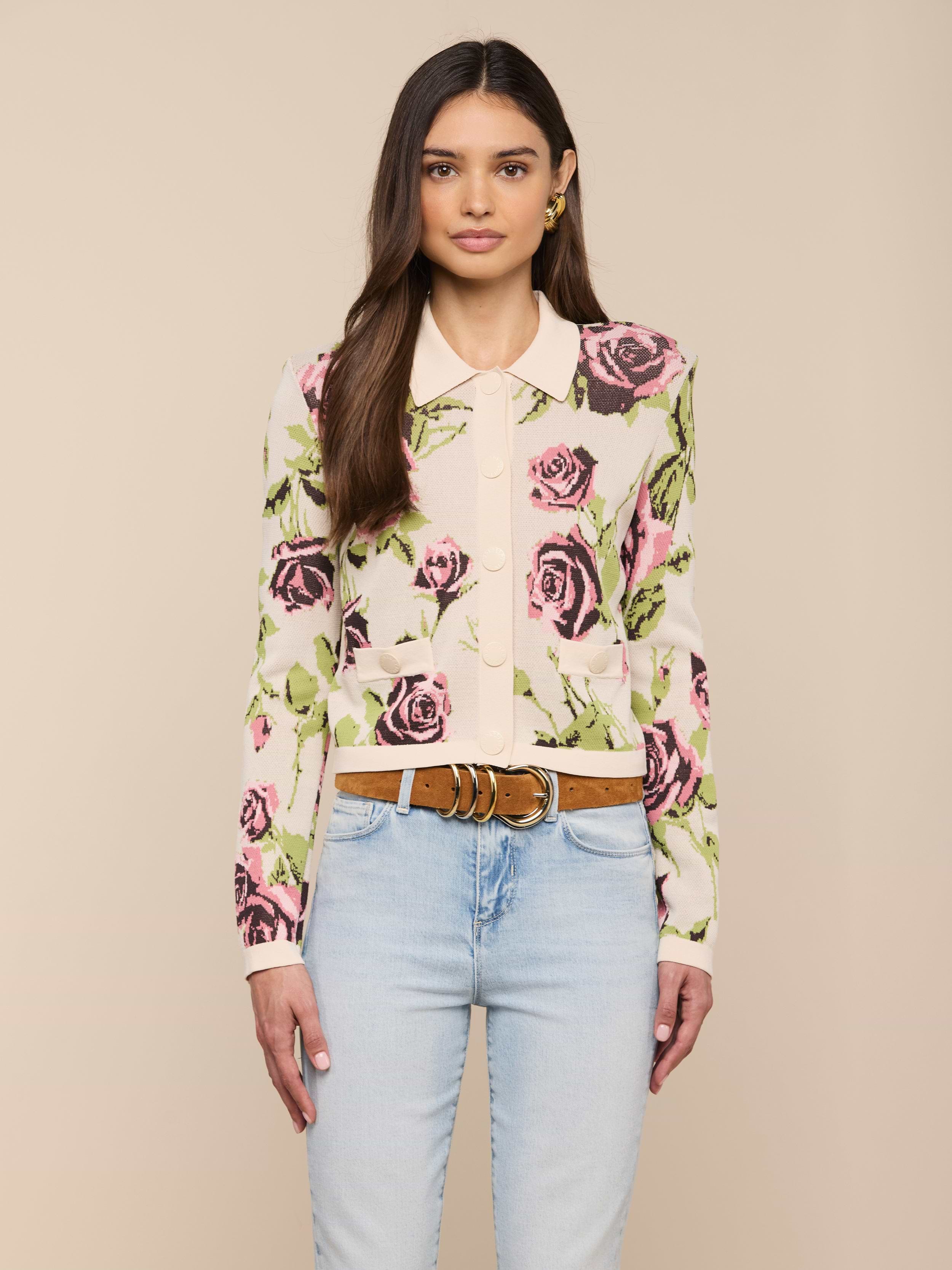 Une femme aux longs cheveux bruns porte le cardigan Osiris Jacquard, avec un grand motif de rose et un col crème, associé à un jean bleu clair et une ceinture marron, sur un fond beige uni.