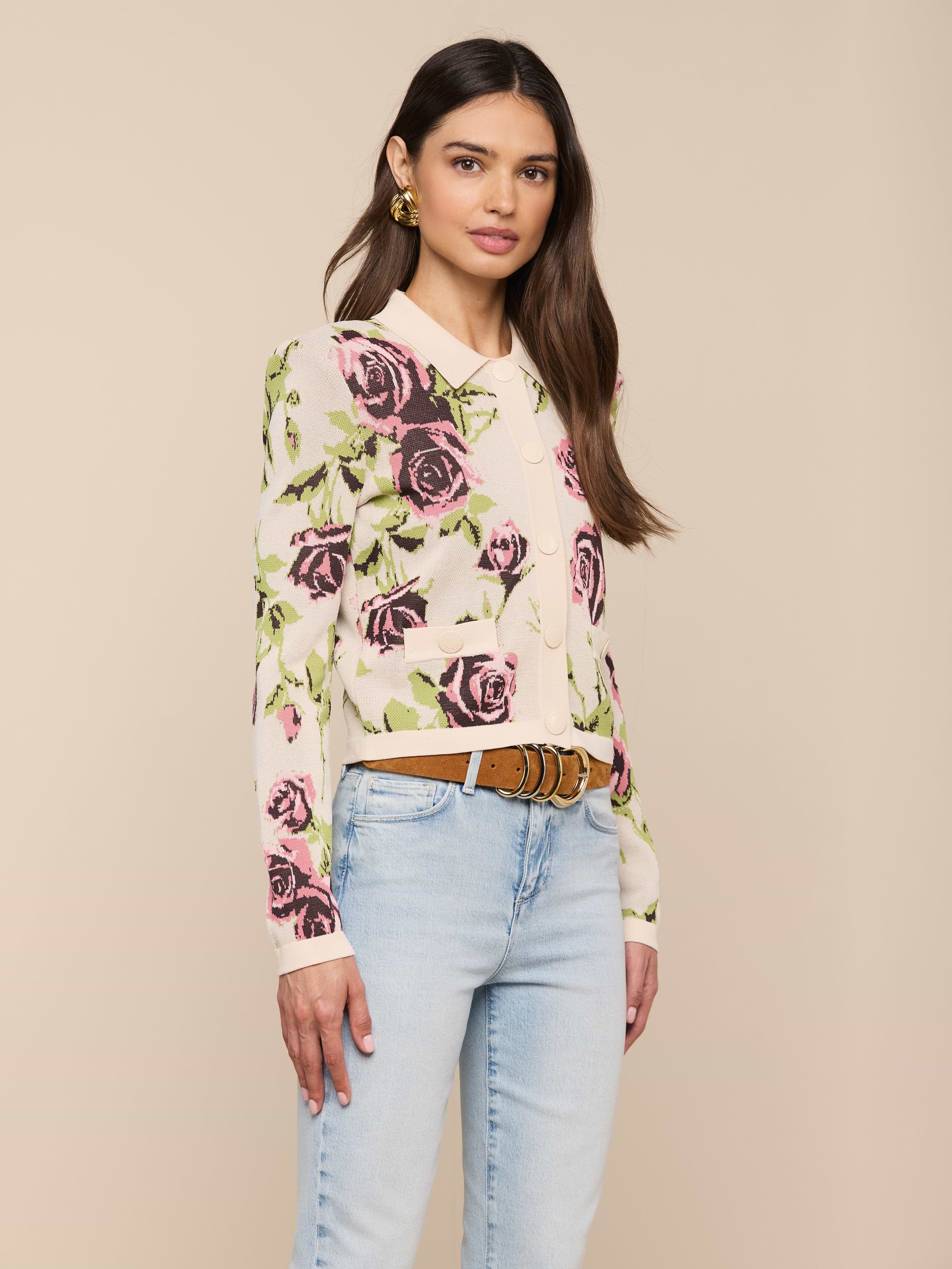 Une femme aux longs cheveux bruns se tient sur un fond beige. Elle porte le cardigan Osiris Jacquard avec un motif de roses roses et violettes, un jean bleu clair, une ceinture marron et des boucles doreilles en or.