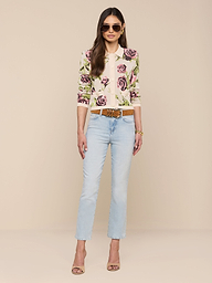 Une femme se tient debout sur un fond beige, portant des lunettes de soleil, des bijoux en or, le cardigan en jacquard Osiris avec un motif de rose, un jean bleu clair avec une ceinture marron et des poches à rabat, ainsi que des sandales à talons hauts.