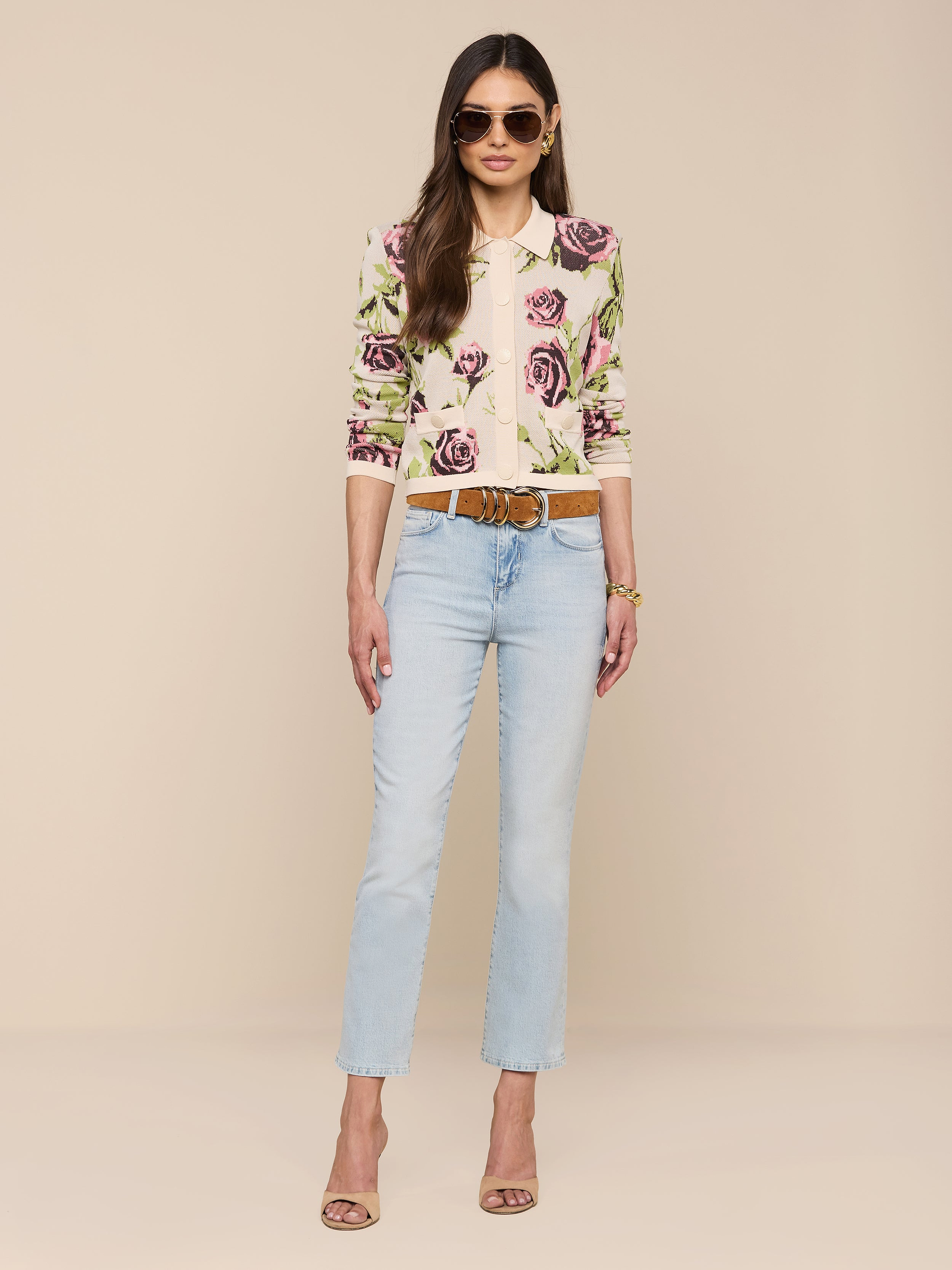 Une femme se tient debout sur un fond beige, portant des lunettes de soleil, des bijoux en or, le cardigan en jacquard Osiris avec un motif de rose, un jean bleu clair avec une ceinture marron et des poches à rabat, ainsi que des sandales à talons hauts.