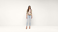 Woman wearing sunglasses, a floral top, light blue jeans, and heeled sandals stands confidently against a plain white background.