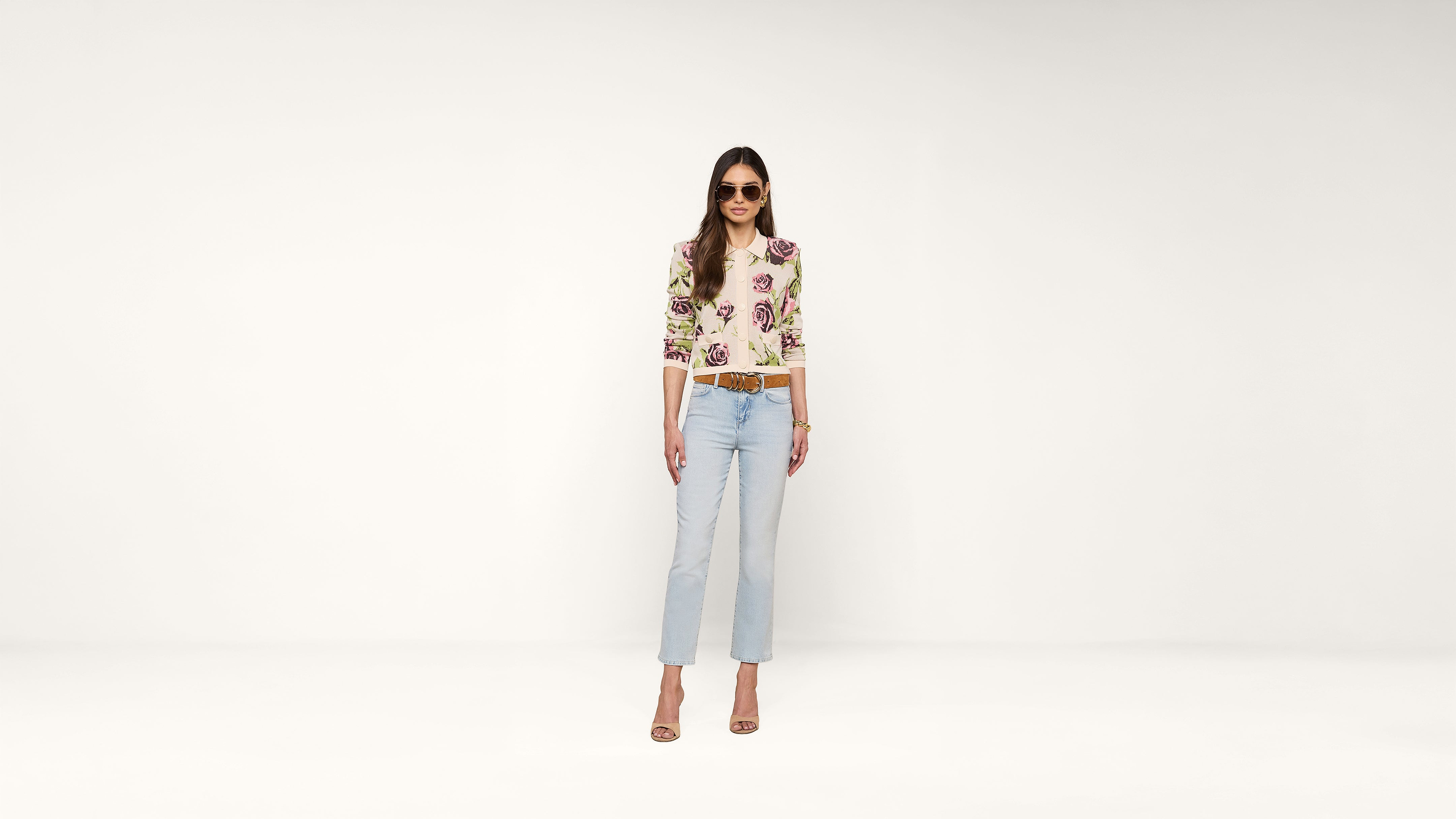 Woman wearing sunglasses, a floral top, light blue jeans, and heeled sandals stands confidently against a plain white background.