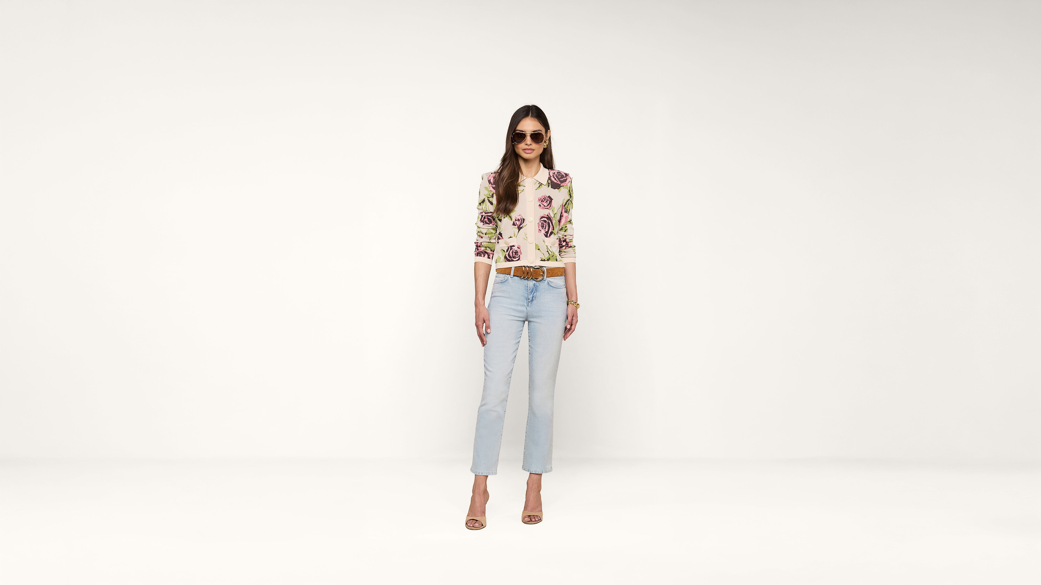 Woman wearing sunglasses, a floral top, light blue jeans, and heeled sandals stands confidently against a plain white background.