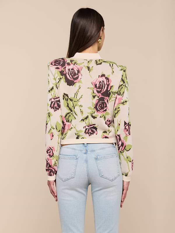 Une femme aux longs cheveux bruns porte le cardigan Osiris Jacquard, avec un motif de roses roses et vertes et des poches à rabat, associé à un jean bleu clair. Elle se tient debout, dos à la caméra, sur un fond beige uni.