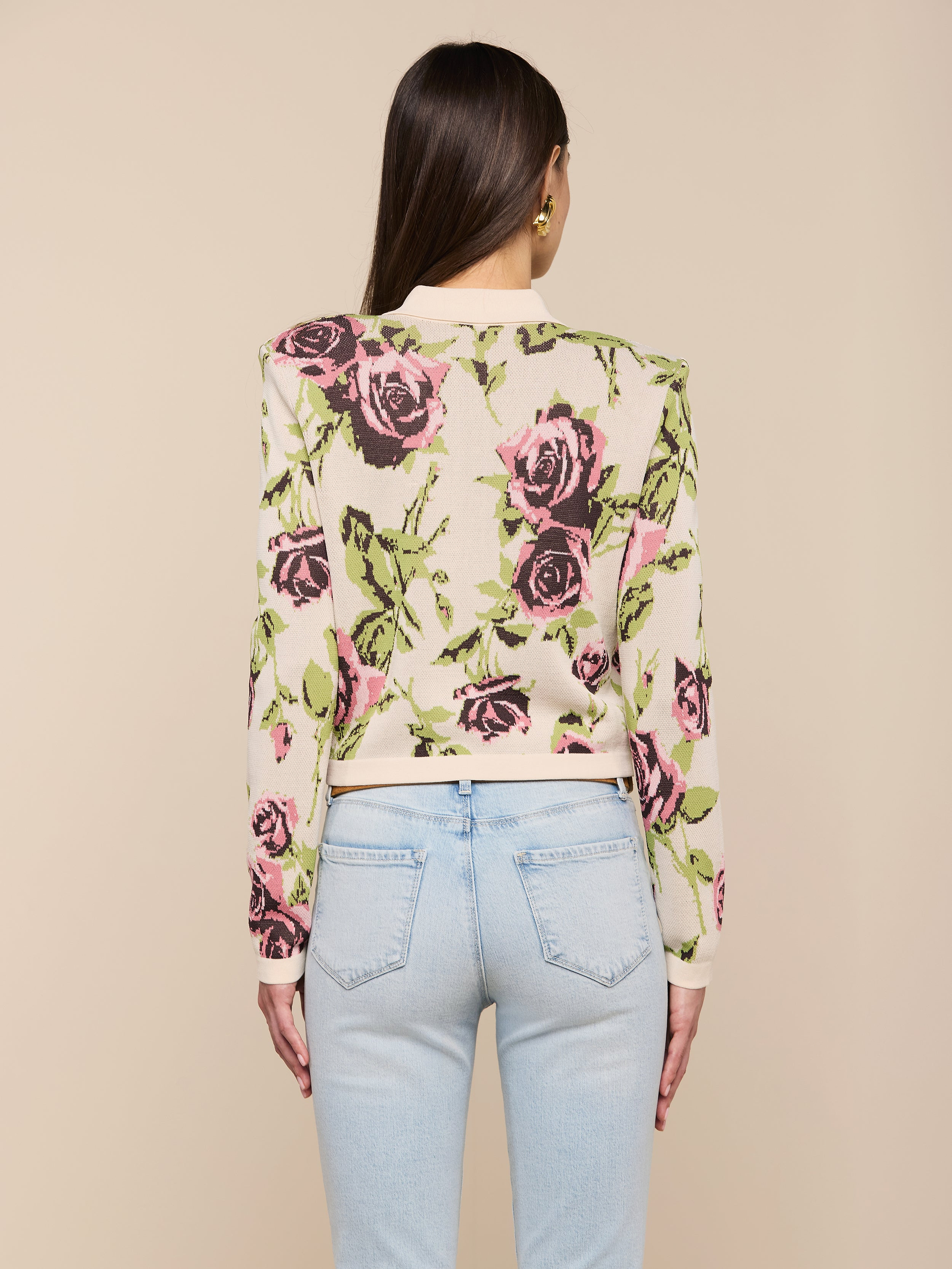 Une femme aux longs cheveux bruns porte le cardigan Osiris Jacquard, avec un motif de roses roses et vertes et des poches à rabat, associé à un jean bleu clair. Elle se tient debout, dos à la caméra, sur un fond beige uni.