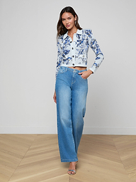 A woman stands wearing the Osiris Jacquard Cardigan, a white jacket with blue floral prints accented by silver buttons, paired with high-waisted wide-leg blue jeans. She has straight hair and is wearing heeled sandals, posing against a plain white background.