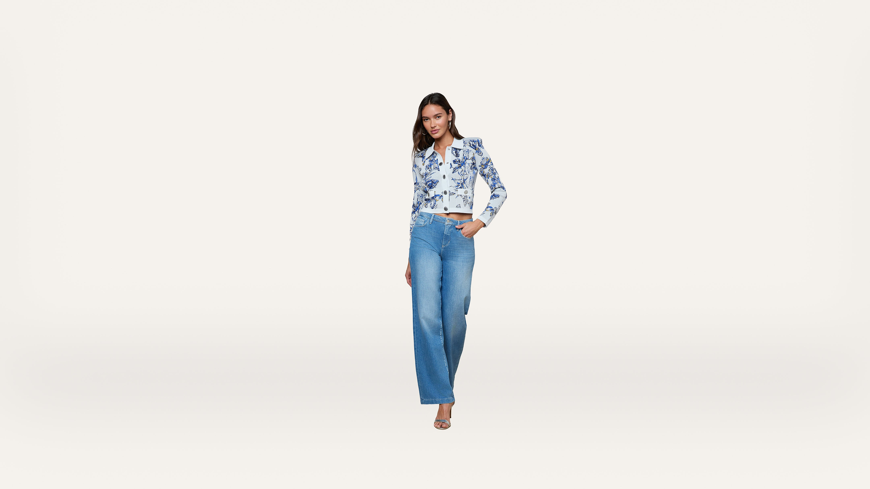 A woman stands confidently in wide-leg blue jeans, a white and blue floral long-sleeve shirt, and heeled sandals against a plain white background.