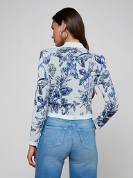 A woman with long brown hair, facing away from the camera with her hands on her hips, wears the Osiris Jacquard Cardigan—a blue and white floral knit jacket with silver buttons—paired with light blue high-waisted jeans, set against a plain light background.