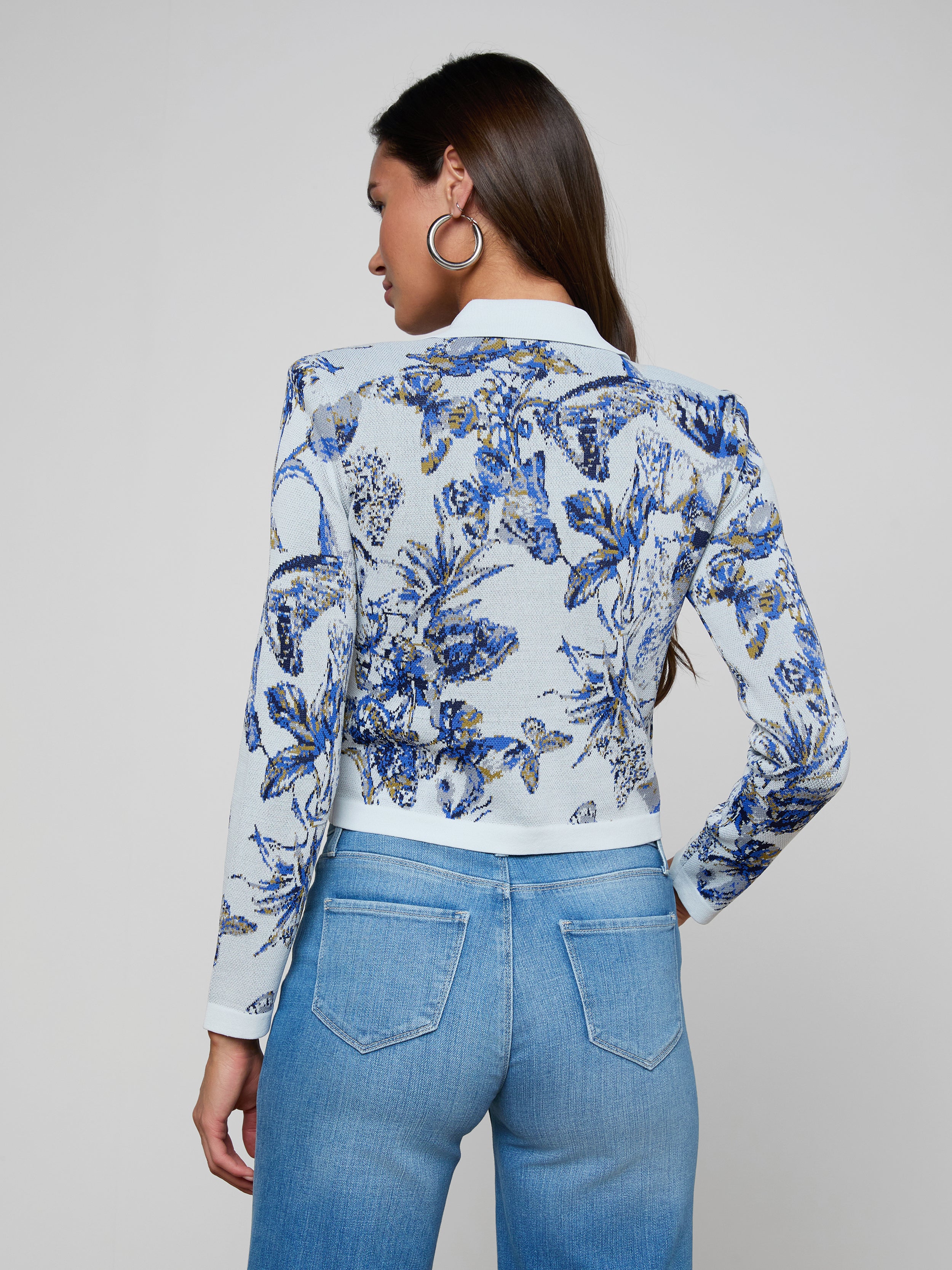A woman with long brown hair, facing away from the camera with her hands on her hips, wears the Osiris Jacquard Cardigan—a blue and white floral knit jacket with silver buttons—paired with light blue high-waisted jeans, set against a plain light background.