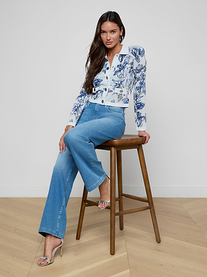 A woman with long brown hair sits on a wooden stool, wearing the Osiris Jacquard Cardigan with blue floral patterns, blue jeans, and silver high-heeled sandals against a white wall and light wooden floor.