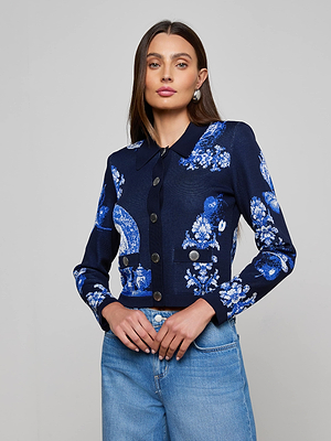 A woman with long brown hair wears the L’AGENCE Osiris Jacquard Cardigan, featuring a navy blue and white floral porcelain motif, paired with high-waisted blue jeans, standing against a plain light gray background.