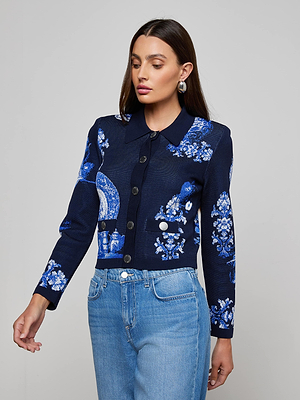A woman models the Osiris Jacquard Cardigan, featuring a blue and white floral pattern, paired with light blue jeans, against a plain gray background as she looks down with one arm slightly extended.