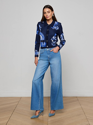 A woman with long brown hair poses indoors against a plain light background wearing the L'AGENCE Osiris Jacquard Cardigan, light blue wide-leg jeans, and blue heels, accessorized with minimal jewelry and one hand in her pocket.