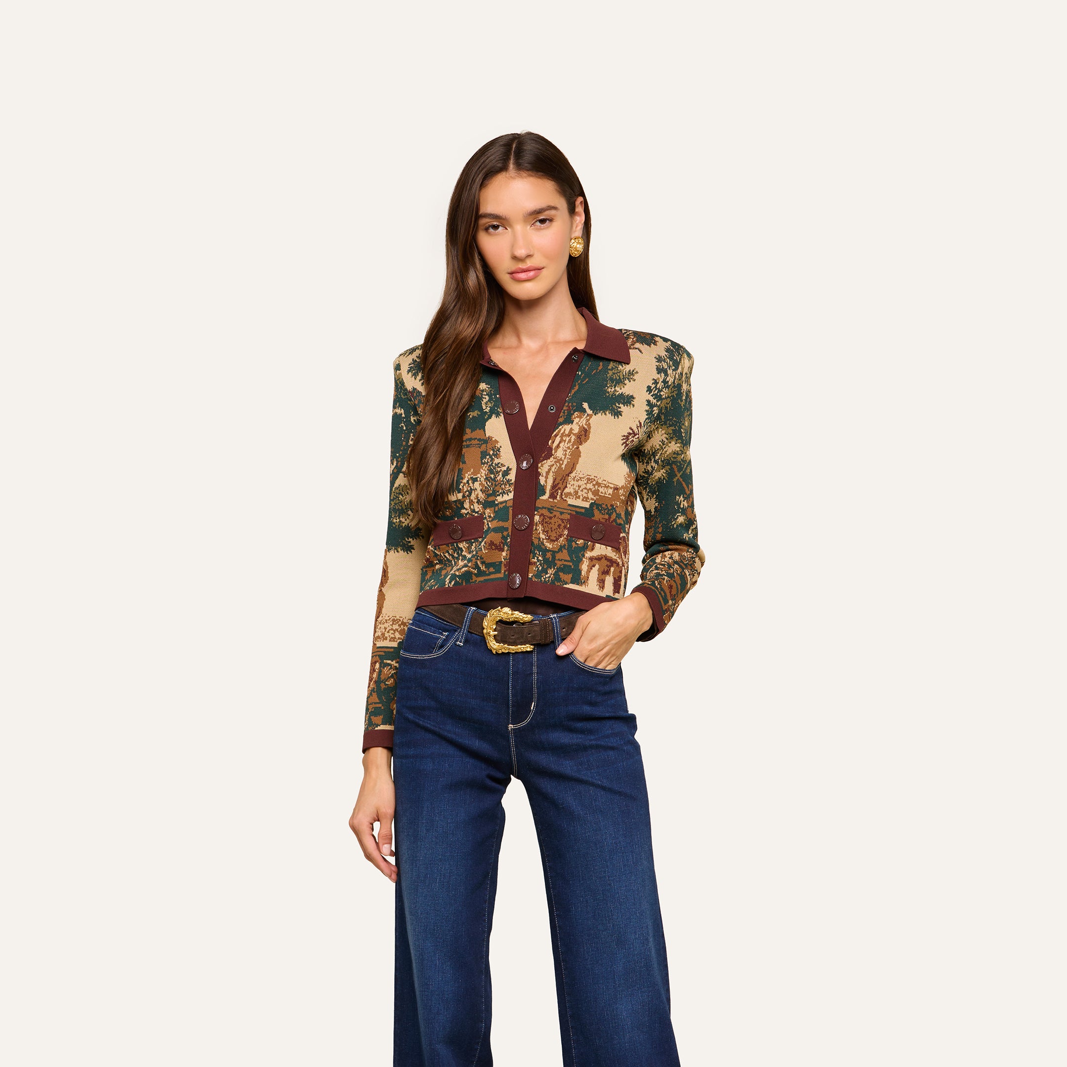 A woman with long brown hair stands against a plain white background, looking at the camera with a relaxed expression. She is wearing the Osiris Jacquard Cardigan, which features a patterned jacquard knit and a brown collar, paired with wide-leg blue jeans.