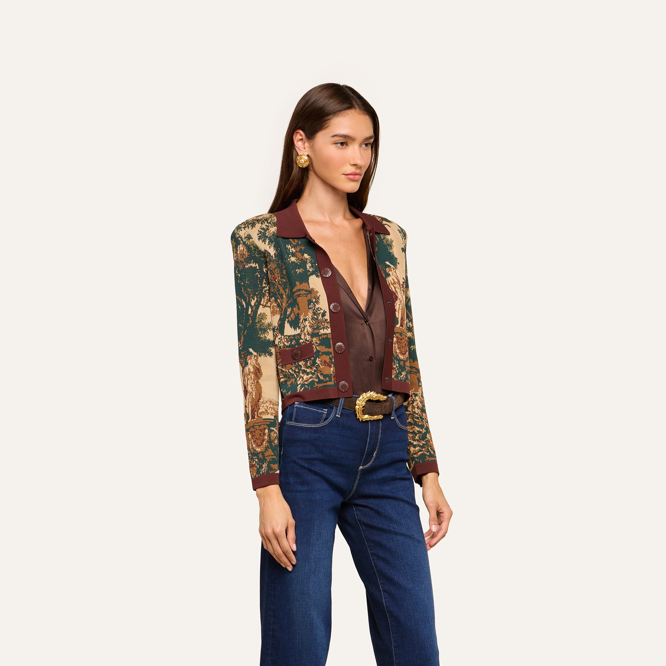A woman with straight brown hair wears the Osiris Jacquard Cardigan, styled over a deep V-neck brown top, blue jeans, and a belt with a decorative buckle, standing against a plain light background.