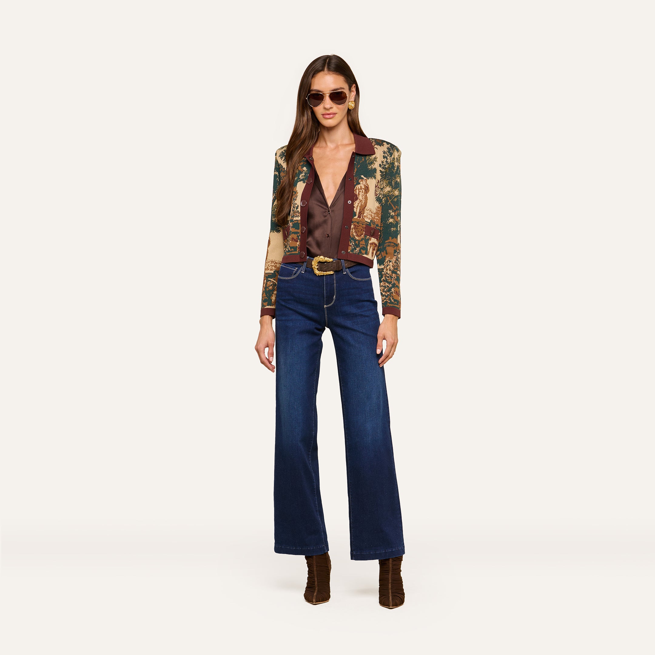 A woman wearing sunglasses, the Osiris Jacquard Cardigan with a tapestry print, a brown blouse, wide-leg blue jeans with a belt, and brown boots stands against a plain light background.