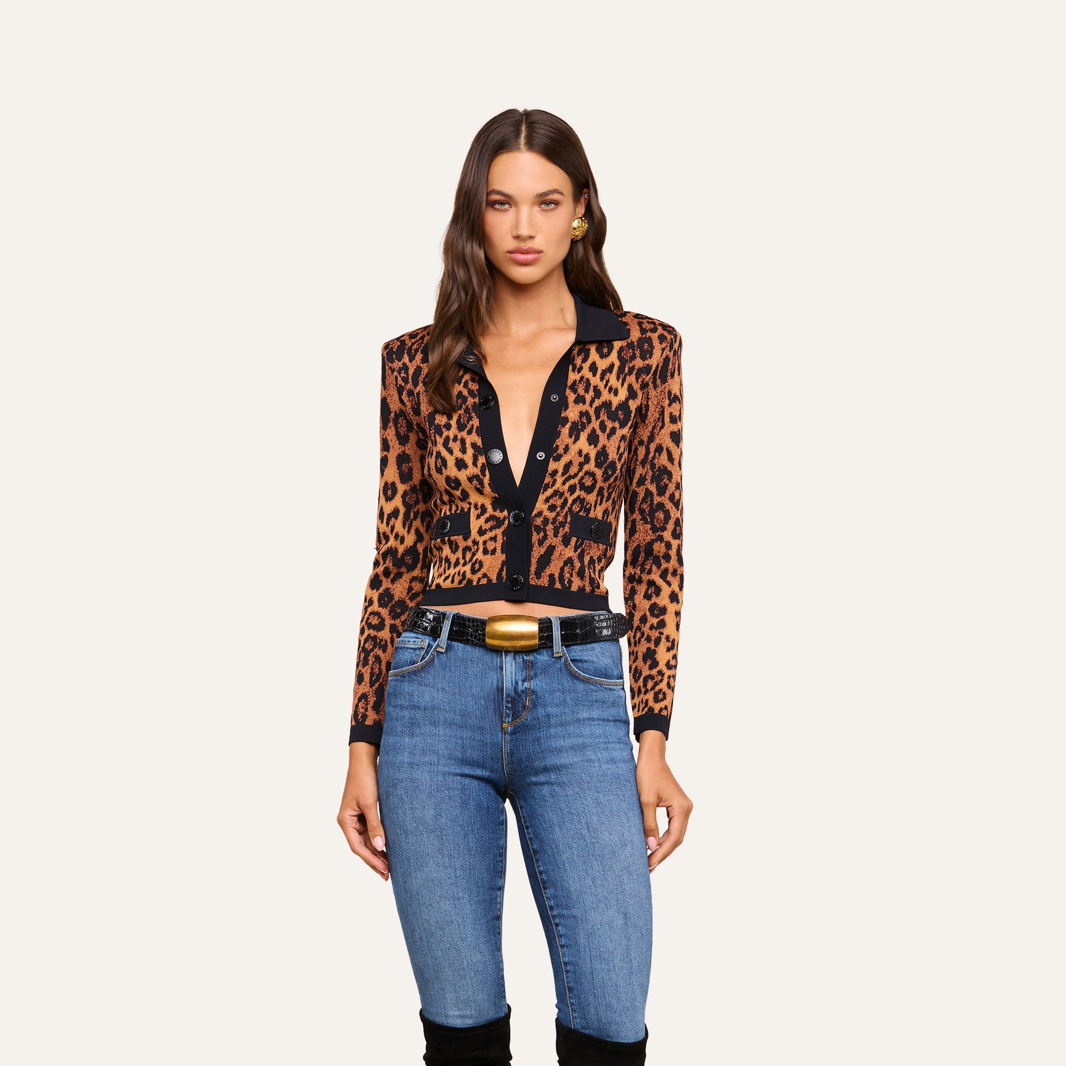 A woman wearing the Osiris Jacquard Cardigan with flap pockets, paired with blue jeans, a black belt featuring a gold buckle, stands against a plain beige background.