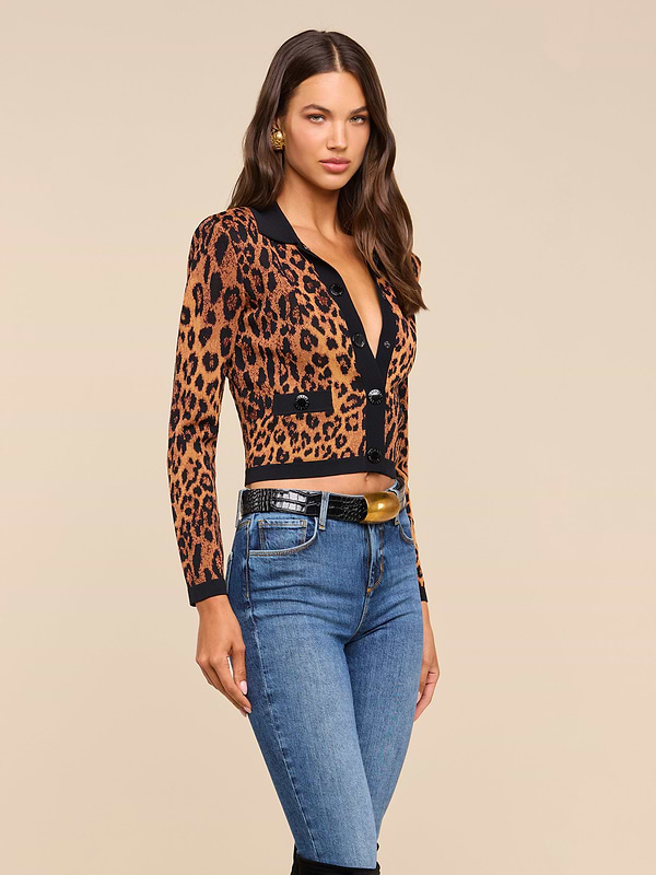 A woman with long brown hair wears the Osiris Jacquard Cardigan—a leopard print knit featuring flap pockets—paired with blue jeans and gold earrings, standing against a plain beige background.