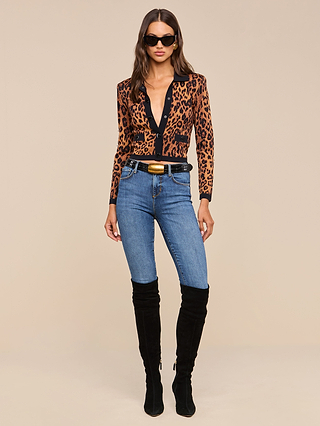 A woman wearing sunglasses and the Osiris Jacquard Cardigan with flap pockets, paired with blue jeans and black knee-high boots, stands against a plain beige background.