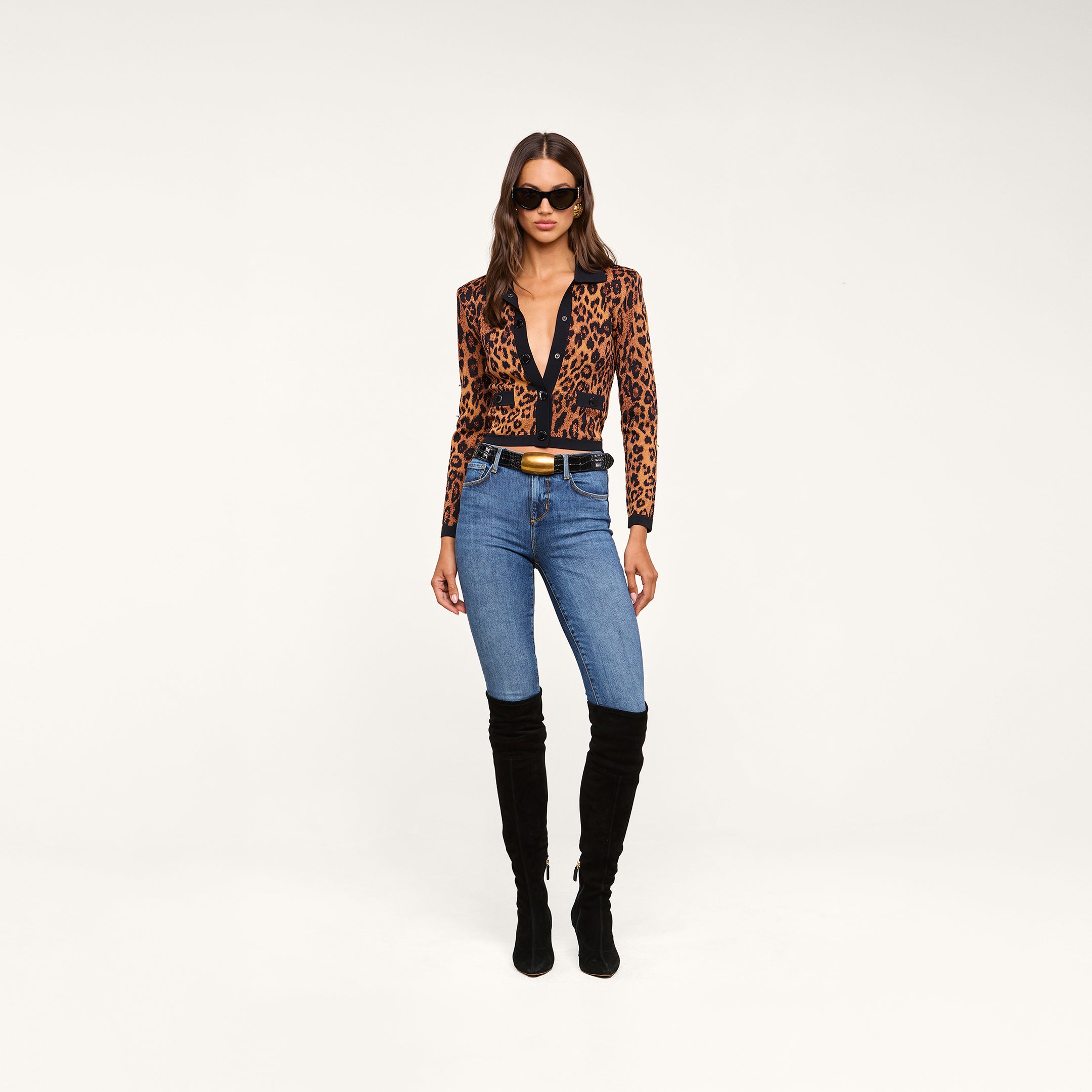 A woman wearing sunglasses and the Osiris Jacquard Cardigan with flap pockets, paired with blue jeans and black knee-high boots, stands against a plain beige background.