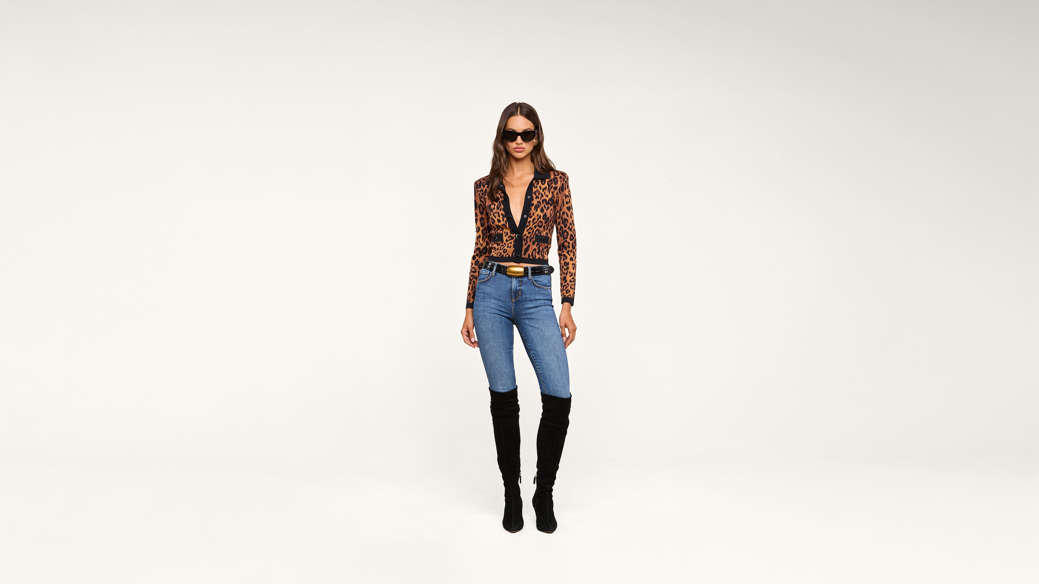 A woman stands against a plain white background, wearing a leopard print top, blue jeans, black knee-high boots, and sunglasses.