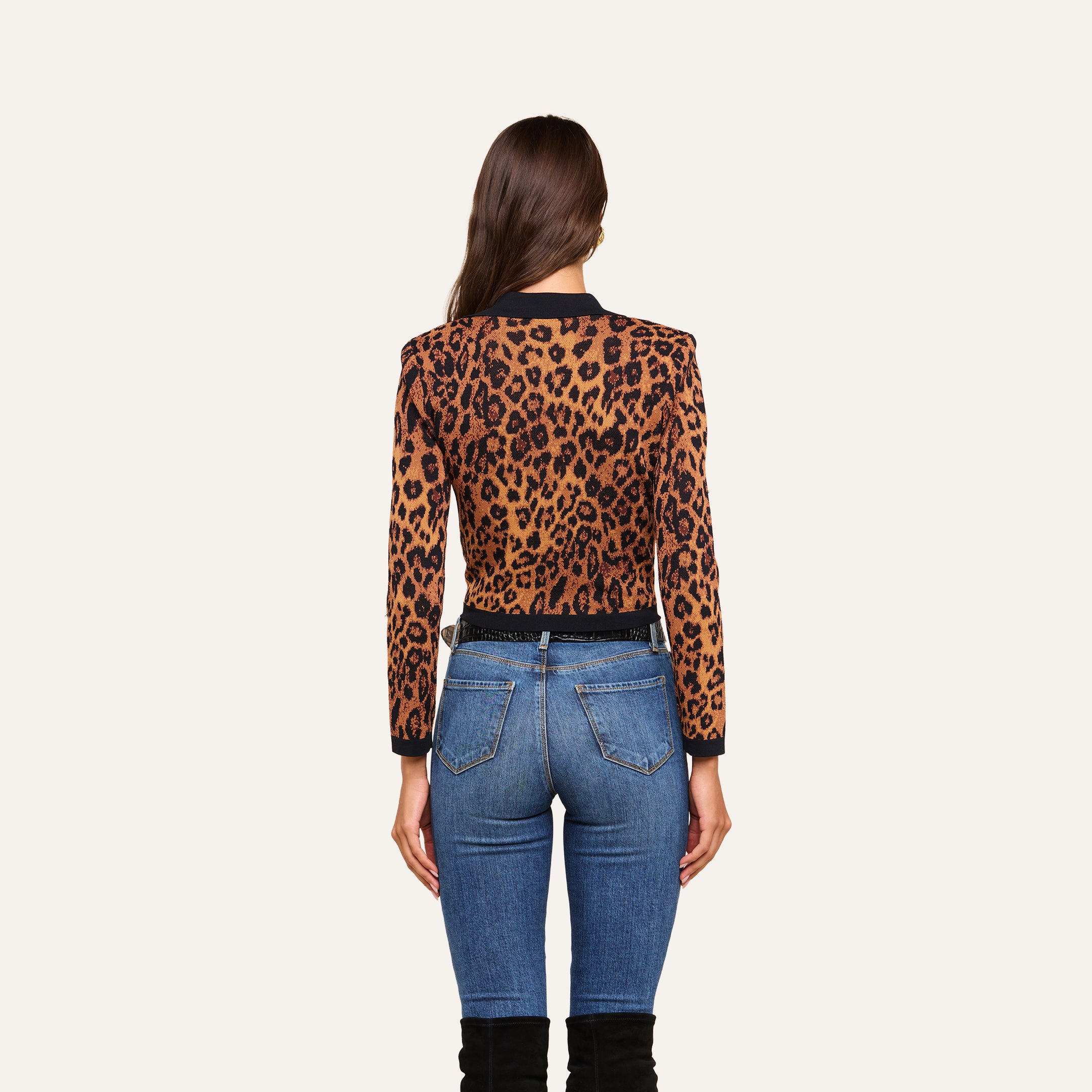 A woman stands facing away against a plain beige background, wearing the Osiris Jacquard Cardigan with flap pockets, blue jeans, and black knee-high boots.