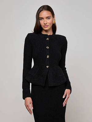 A woman with long brown hair wears L'AGENCE's Melissa Knit Cardigan—black, textured, with gold buttons and a sculpted waist—paired with a matching skirt and gold earrings, standing before a plain light gray background.