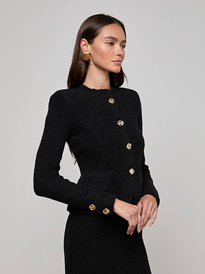A woman with long brown hair wears the Melissa Knit Cardigan by L'AGENCE, featuring textured knit and gold buttons, paired with a matching skirt. She stands sideways with folded hands, large gold earrings brightening a plain light background.