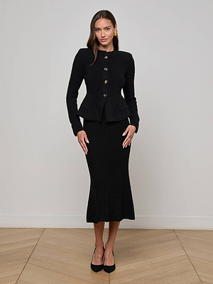 A woman stands indoors on a wooden floor, wearing the fitted black Melissa Knit Cardigan by L'AGENCE with gold buttons and a matching calf-length skirt. She pairs it with gold earrings, black heels, and has long hair against a light wall.
