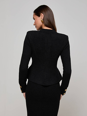 A woman with straight brown hair, wearing the L'AGENCE Melissa Knit Cardigan in black with gold buttons, stands facing away from the camera. She accessorizes with gold earrings against a plain light gray background.