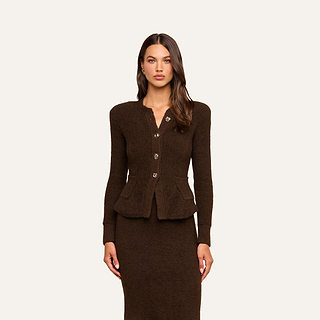 A woman stands facing forward, wearing the Melissa Knit Cardigan—an elegant, fitted brown design crafted from textured yarn with gold buttons—paired with a matching skirt, set against a plain beige background.